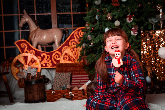 Funny happy little girl child in pajamas sitting in christmas decorated room and licking a caramel candy in the shape of a snowman. New Year's eve. St. Nicholas Day. Present. Time of holidays.	
