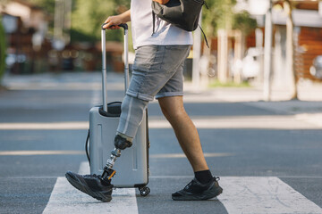 Man with disability, with a prosthetic leg, walking across a pedestrian crossing carrying a backpack and pushing a wheeled suitcase.