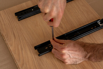 Hands screwing black metal rail to wooden board, close-up, warm light. Male hands using screwdriver to assemble drawer guide on oak surface