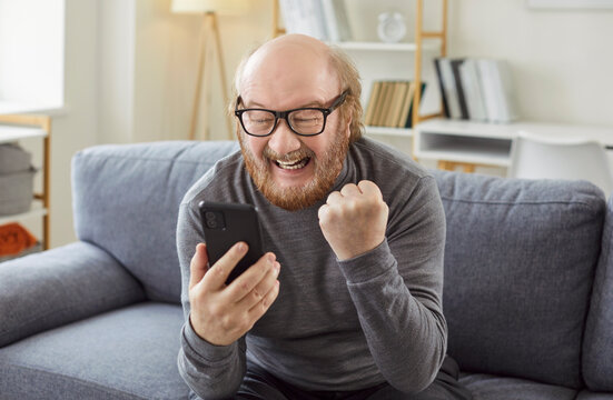 Win money online. Happy emotional elderly man on couch gestures fist pump after social media giveaway surprise award, sale cashback, or web entertainment bookmaker wager victory notification on phone
