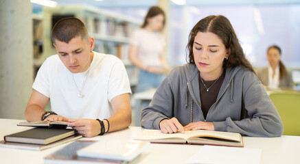 European guy and a girl study together in the library, read books and prepare for university. Students are looking for materials for a lecture in the library of the institute