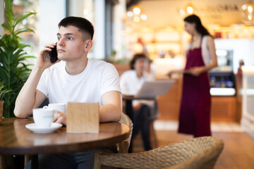 Guy in cafe drinks coffee and using mobile phone, chats online, calls friend, talks on phone. Free Wi-Fi zone, Internet cafe.