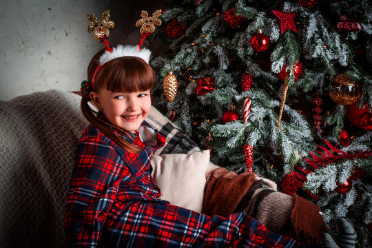 Happy funny little girl child in pajamas wearing a carnival hoop on your head smiling sitting near Christmas tree. New Year's party. Winter holidays eve.