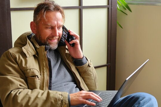 Smiling man working on laptop indoors.Middle-aged man sitting indoors, talking on smartphone and working on a laptop, wearing casual clothes and a warm jacket, natural light from window.