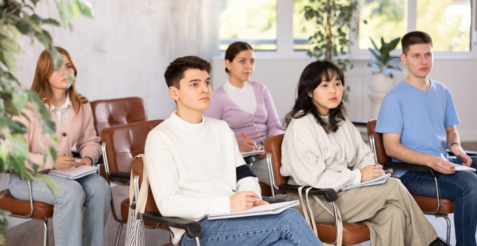 Group of grown-up students attending lecture interestedly and taking notes in light classroom