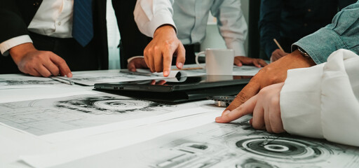 A diverse group of professionals engaged in a business meeting, analyzing technical drawings and discussing plans around a workspace table with laptops and documents. SACTR
