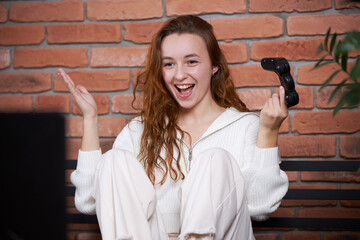 A young beautiful girl sits on the sofa holding a gamepad in her hands and playing a computer game. The player rejoices in victory