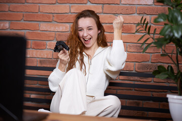 A young beautiful girl sits on the sofa holding a gamepad in her hands and playing a computer game. The player rejoices in victory