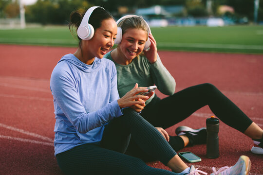 Two diverse female friends and athletes, one Asian and one Caucasian, happily share music and look at a mobile phone during a relaxed break on a running track