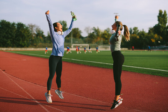 Energetic diverse female athletes celebrate a fitness success, jumping for joy on a running track and raising their water or sport drink bottles in triumph