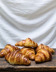 Freshly baked golden croissants stacked on rustic wooden board, flaky buttery pastries with soft fabric background, artisan bakery style