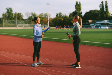 Two diverse female friends and athletes, one Asian and one Caucasian, stand and chat enthusiastically on a running track while taking a hydration break after a workout