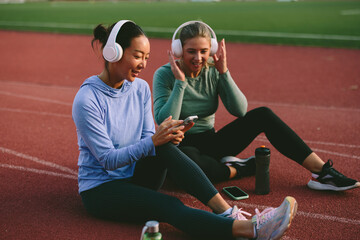 Two diverse female friends and athletes, one Asian and one Caucasian, happily share and view content on a mobile phone while wearing headphones during a fitness break on a running track