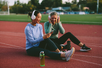 Two diverse female friends and athletes, one Asian and one Caucasian, happily listen to music on shared headphones and look at a mobile phone during a fitness break on a running track