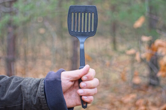 A man's hand holds one black large plastic slotted spoon on an autumn street
