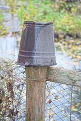 One old brown enameled metal bucket hangs on a wooden post of an old fence near the water of a lake on a summer street