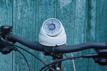 One white webcam stands on a black bicycle handlebar near a green wall on a summer street