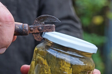 A hand with an old rusty can opener opens a white metal lid on a glass jar with green pickled cucumbers