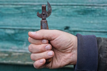 One hand of a man holding an old iron rusty brown can opener against the background of a green wall on the street