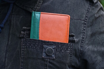 A notebook with a red and green leather cover lies in the pocket of a gray and black jacket on a man on the street