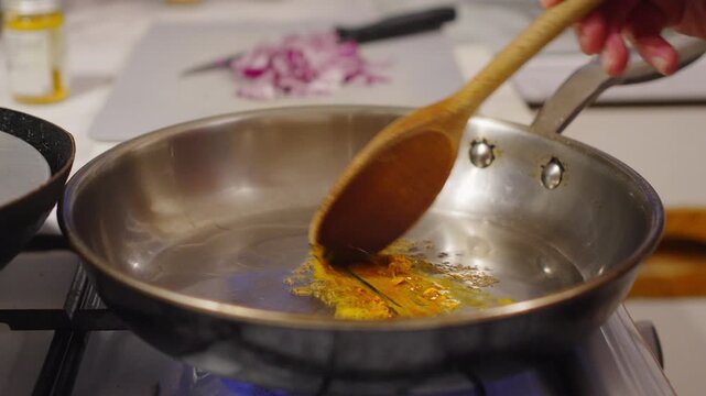 Spices sizzling in oil as a wooden spatula stirs; chopped onions await on the board nearby in home kitchen