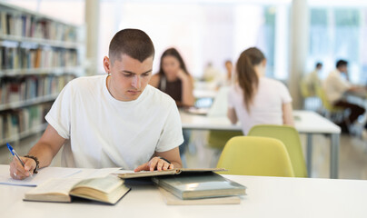 Male student sits at table in library and works with books, printed literature, writes out theses and records key points, preparation for exam....