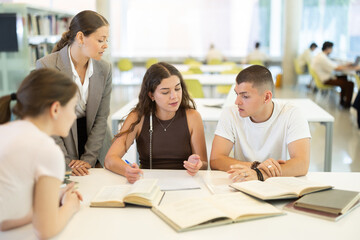 Group of classmates sits at table in library and works with books and printed literature, writes out theses and records key points, deliberative meeting, collective work, preparation for exam