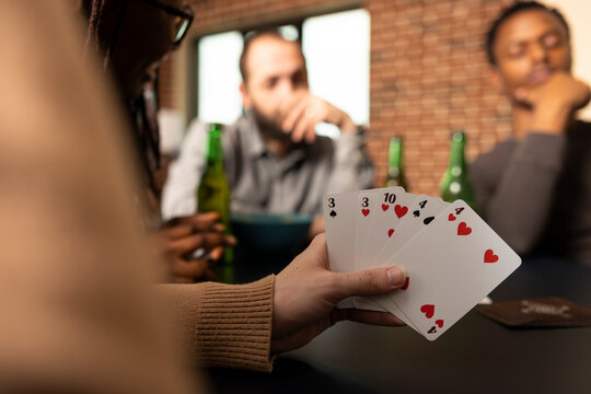 Selective focus on caucasian woman grasping poker cards during game day with diverse friends at home. Multiethnic group sits around living room table enjoying fun leisure activity in brick wall room.