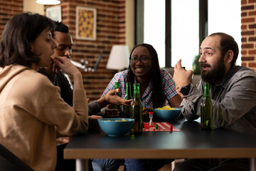 Young adults pause from board game to listen intently as white man shares an engaging story. Diverse friends gathered around the table with alcoholic beverage and bowl of chips, hanging out indoors.