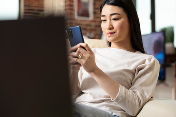 Close up of asian business advisor in casual outfit sitting on sofa, relaxing during remote work. Female freelancer browsing social media on smartphone and chatting with friends.