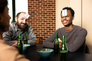 African american and caucasian men sit with cards on foreheads, surrounded by snacks and drinks during hangout. Laughter breaks out as friends gather at table, enjoying lighthearted guessing game.