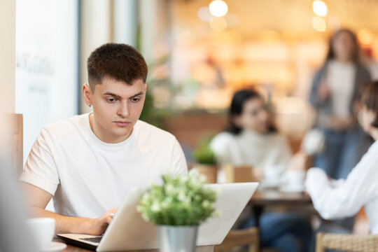 Young man is sitting at a table in a cafe and working at a laptop. Guy designer works remotely in a coffee shop - Powered by Adobe