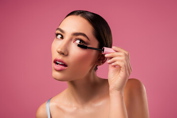Beautiful young woman applying mascara against a vibrant pink background, showcasing elegance and a sleek
