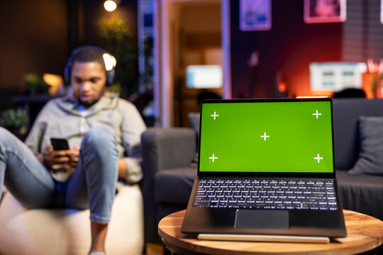 Black young person listening to music on headset next to green screen, feeling casual and relaxed on the bean bag in his living room. Happy chill man in his trendy loft with gadgets.