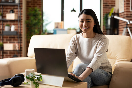 Cheerful female entrepreneur sitting confidently on sofa with laptop in home office. Smiling businesswoman working remotely, ready to review online articles to boost company progress. - Powered by Adobe