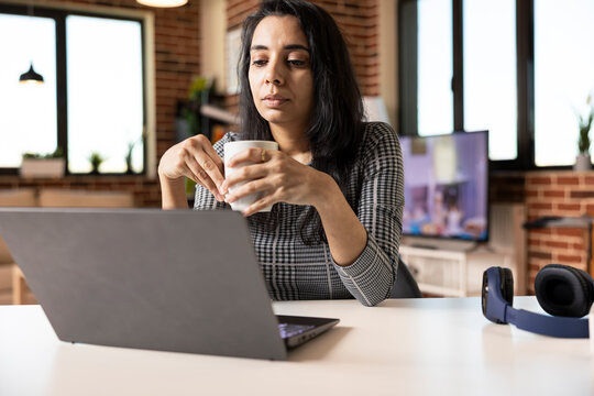 Professional indian woman seated at desk holding tea mug while focused on laptop screen. Remote female employee learning new business skills during online course or webinar from home office.