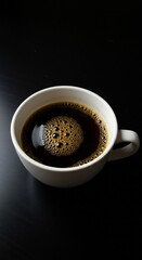 A close-up view of a white coffee cup filled with dark coffee, showcasing the surface with bubbles. The background is a dark surface, enhancing the contrast.