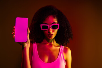Portrait of stylish young woman in neon lighting wearing sunglasses and posing with a smartphone, modern trendy vibes