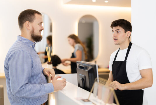 Young guy hairdresser stylist at cash register consulting young male client in beauty salon