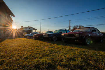 Classic cars are parked in a grassy yard at sunset, their silhouettes catching the warm golden light. A quiet rural setting with houses and power lines forms a calm backdrop to the vintage lineup.q