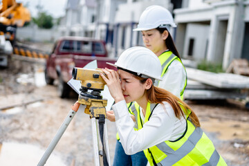 Two female engineers using theodolite for land survey at housing construction site, wearing reflective safety vests and hard hats while checking blueprints and project measurements