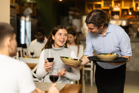 Woman, sitting with her husband at the table, drinks wine. Male waiter brings cooked food for a couple in a restaurant.