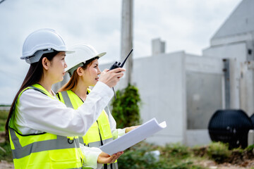 Two female engineers wearing safety helmets and reflective vests inspecting construction site and discussing project plan with blueprint, real estate developer, construction industry.