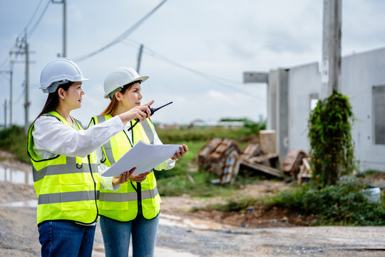 Two female engineers wearing safety helmets and reflective vests inspecting construction site and discussing project plan with blueprint, real estate developer, construction industry. - Powered by Adobe
