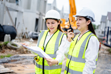 Two female engineers wearing safety vests and helmets discussing construction blueprints at residential building site with crane in background, teamwork and project planning concept
