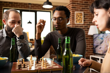 Multiethnic group of friends seated around table, enjoying a board game with snacks. African american man with chess piece in hand, looking at chessboard on table along with his caucasian companions.