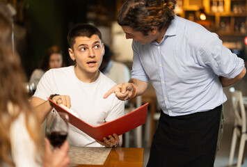 Male waiter tells a restaurant client in detail about the dishes, food and the concept of the...