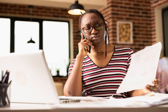 Confused remote worker sits at cluttered table, frustrated by incomplete paperwork while handling business issues via smartphone. Businesswoman visibly upset during tense conversation with colleague.