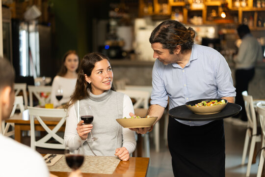 Woman, sitting with her husband at the table, drinks wine. Male waiter brings cooked food for a couple in a restaurant.