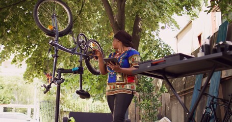 Dedicated african american lady using phone tablet to research for solutions of damaged bike tire in yard. Sports-loving black woman inspecting and repairing bicycle with device and expert work tool.